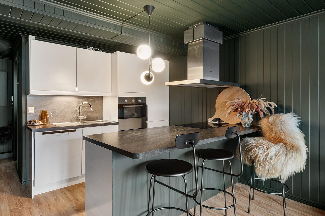 Cocina moderna con mobiliario blanco y paredes en tono verde oscuro, equipada con isla central de superficie negra, taburetes altos, lámparas de diseño contemporáneo y detalles decorativos como un florero y una manta de pelo sobre la silla.