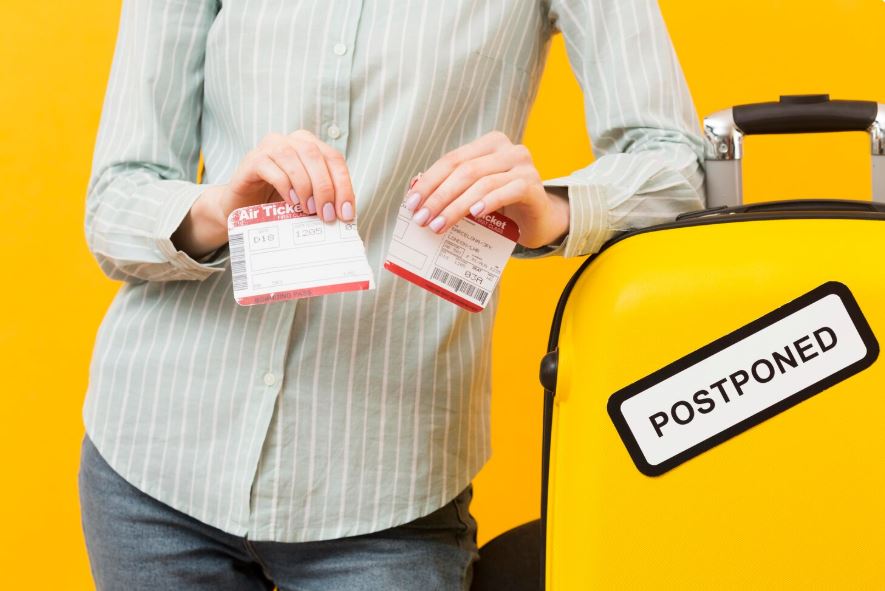 Persona sujetando billetes de avión rotos junto a una maleta amarilla con cartel de vuelo pospuesto.