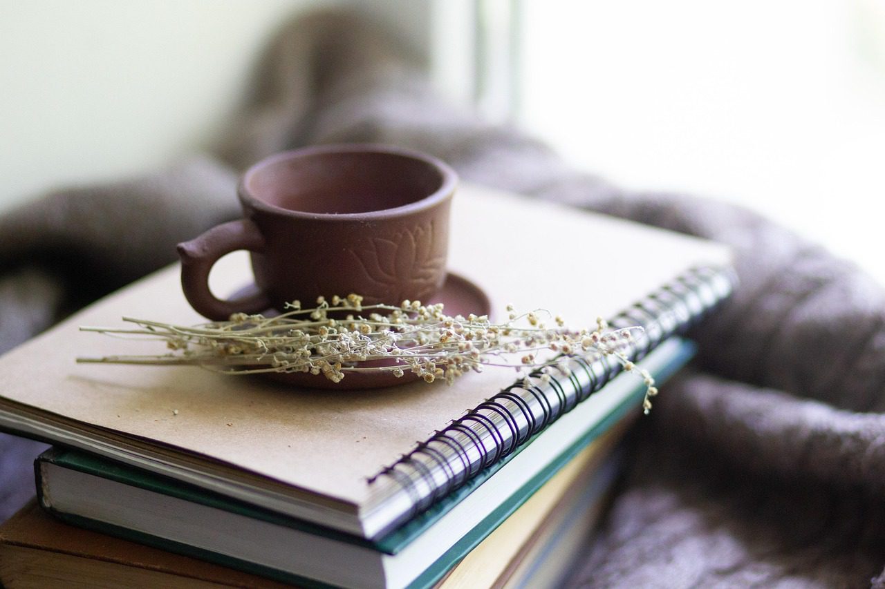 Taza de cerámica con infusión de hierbas secas sobre un cuaderno de espiral y una pila de libros, en un ambiente cálido de lectura y bienestar.