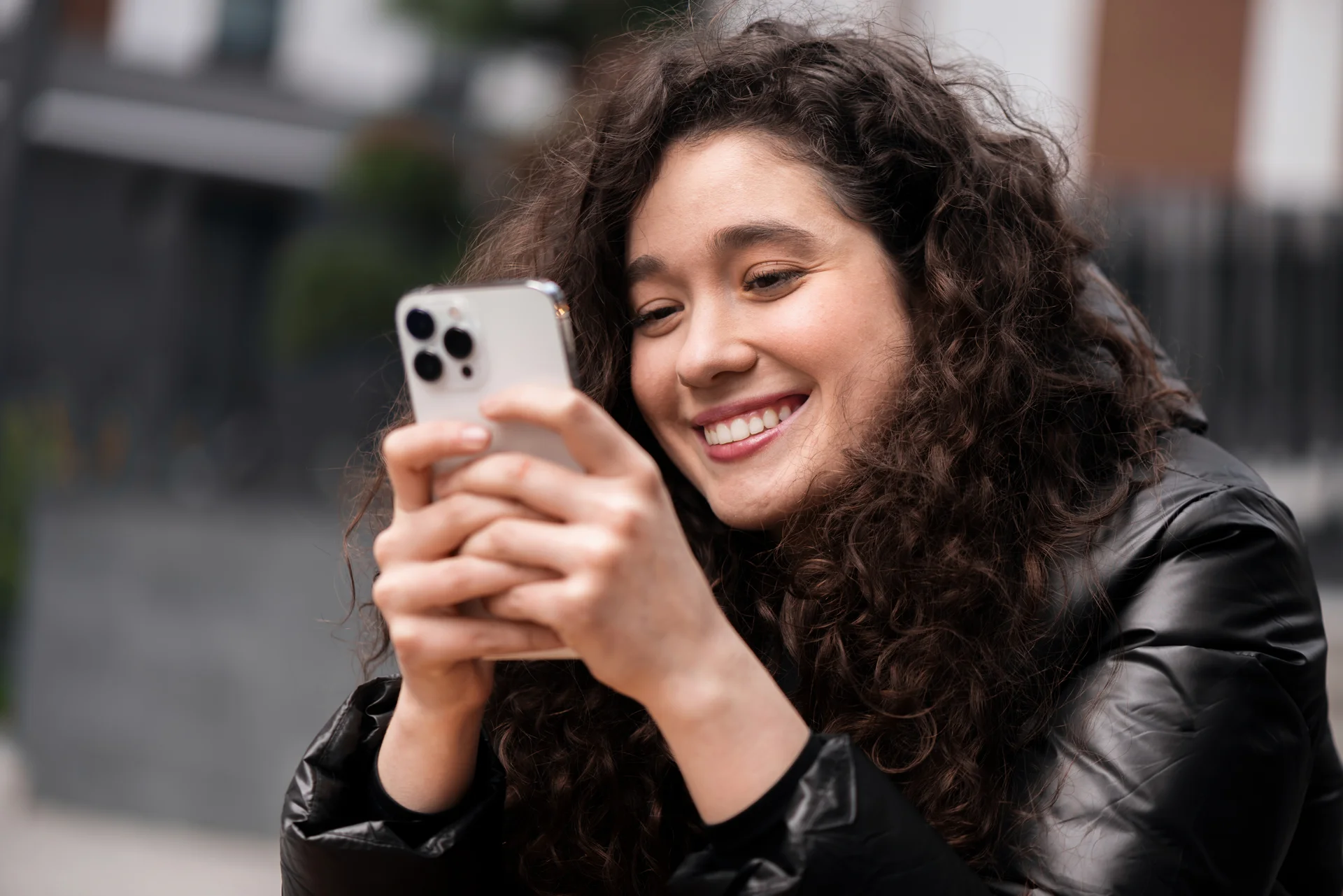 Mujer joven sonriente usando un smartphone con ambas manos mientras consulta la pantalla en un entorno urbano